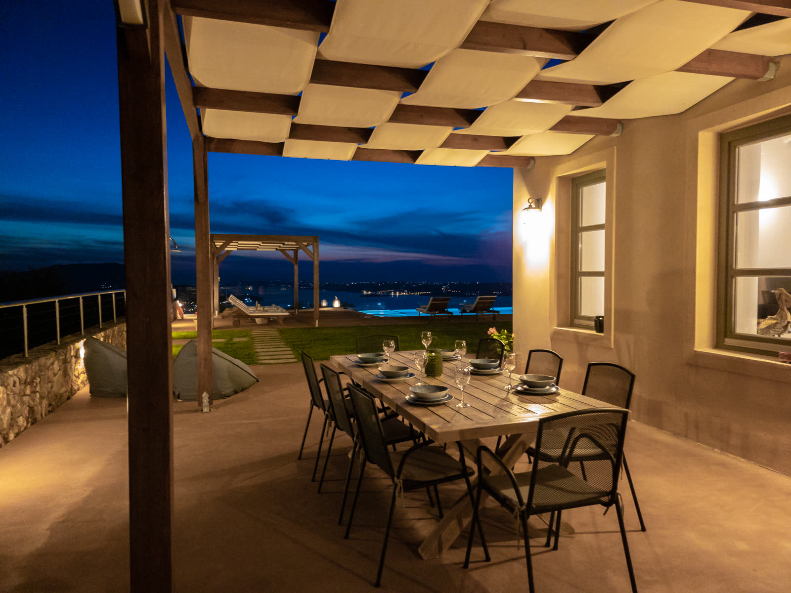Outdoor dining table set under a pergola at twilight, with sea and village lights beyond